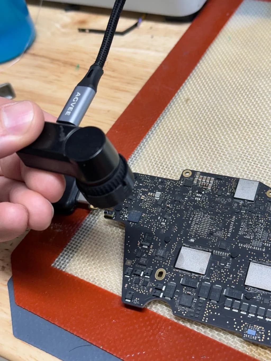 Underside of a 16-inch MacBook Pro A2991 (M3 Pro / M3 Max) logic board secured on a silicone rework mat at Computer Clinic Irvine, being preheated with a hot air rework station for leaky capacitor and MOSFET replacement during microsoldering repair