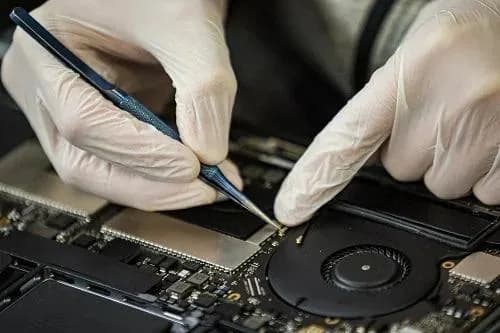 Technician at Computer Clinic Irvine performing board-level microsoldering on a 16-inch MacBook Pro A2991 (M3 Pro / M3 Max) logic board using a hot air rework station and precision tweezers to repair a high current draw fault